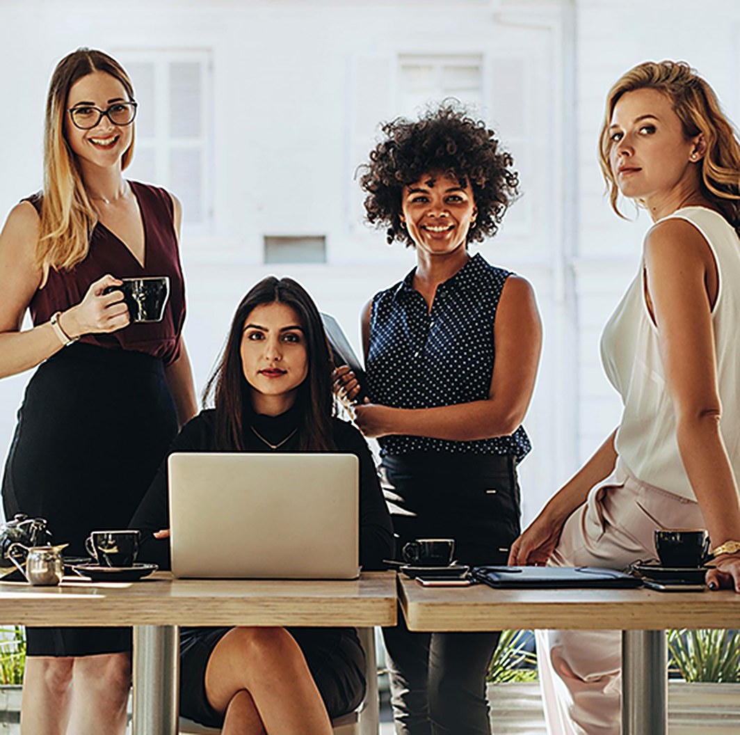 Four women professionals gathered around a table with laptops and coffee, smiling